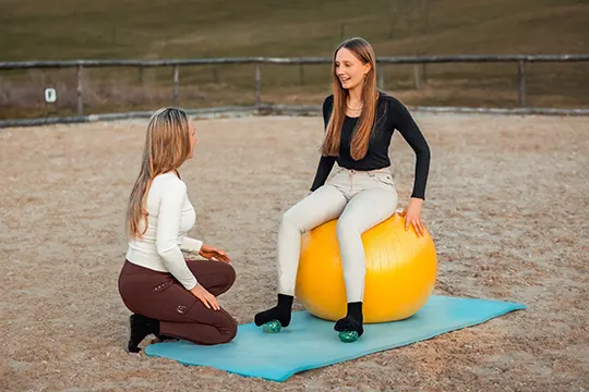 Eine junge Frau sitzt auf dem Reitplatz auf einem großen gelben Ball. Eine andere Frau kniet vor ihr uns zeigt ihr die korrekte Fußstellung.